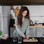 hands holding a glass of water near a cutting board with carrots and leafy greens